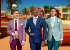 Three men walking outdoors in stylish summer suits, wearing lightweight tailored blazers and trousers in pastel and blue tones, set in a warm resort-style location with palm trees and bright sunlight.